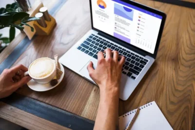 Imagem de um laptop exibindo gráficos e dados enquanto uma pessoa toma café em uma mesa. Perfeito para entender o passo a passo para criar campanhas de Google Ads que convertem.