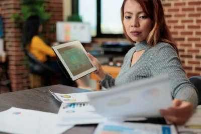 Mulher de cabelo vermelho analisando dados em tablet e documentos em ambiente de escritório, ilustrando o uso de Marketing Preditivo para avançar no marketing de dados